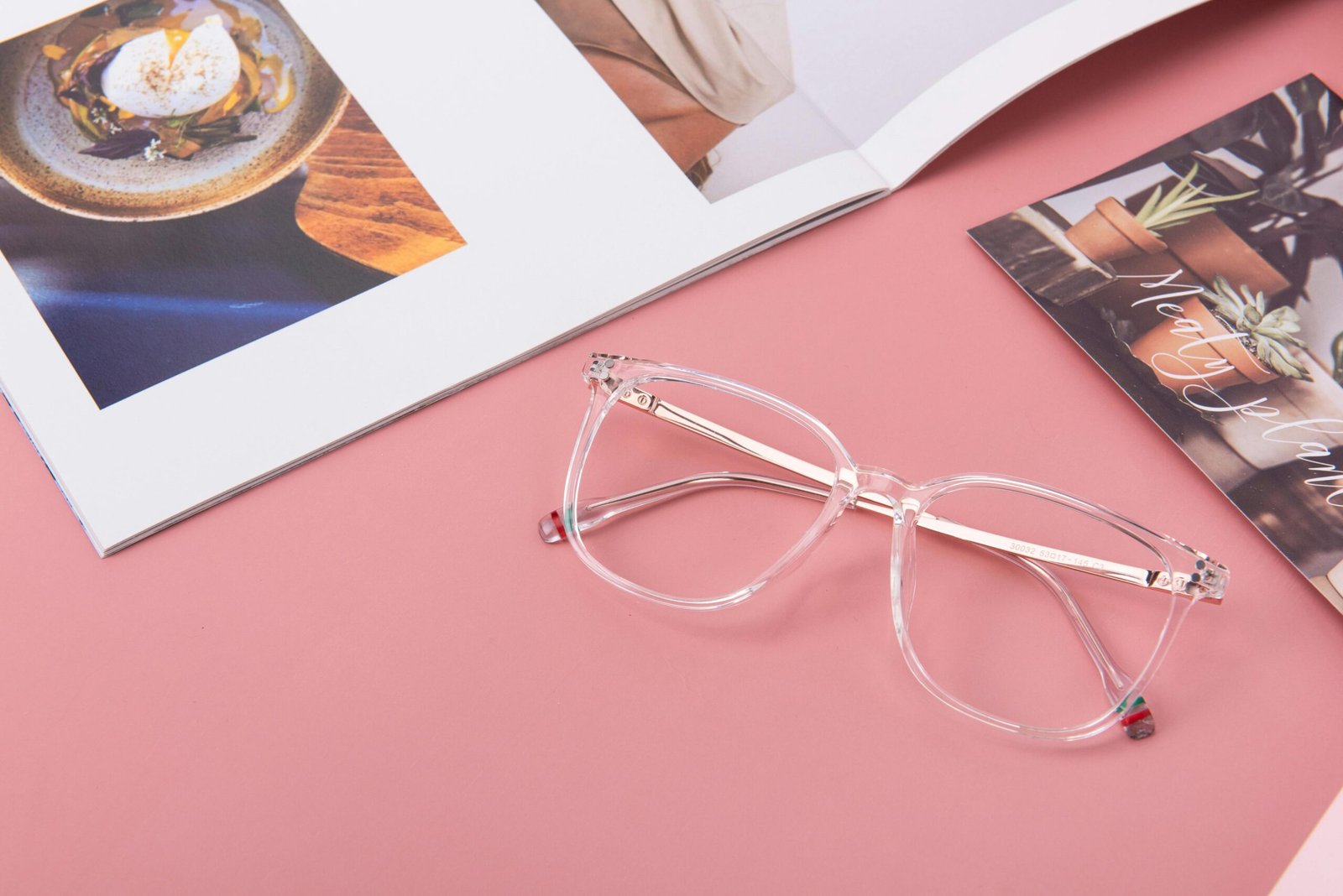 Clear eyeglasses resting on a pink surface with an open book and a plant-themed card.